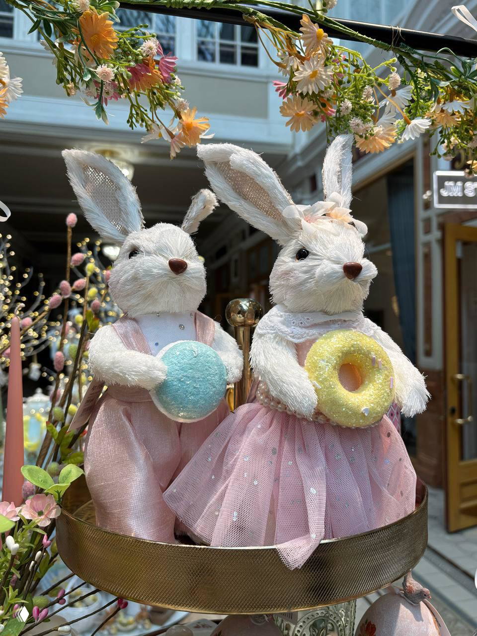 Крупный план пасхальных декоративных зайцев в платьях, держащих яркие аксессуары — часть экспозиции ярмарки в «Пассаж»