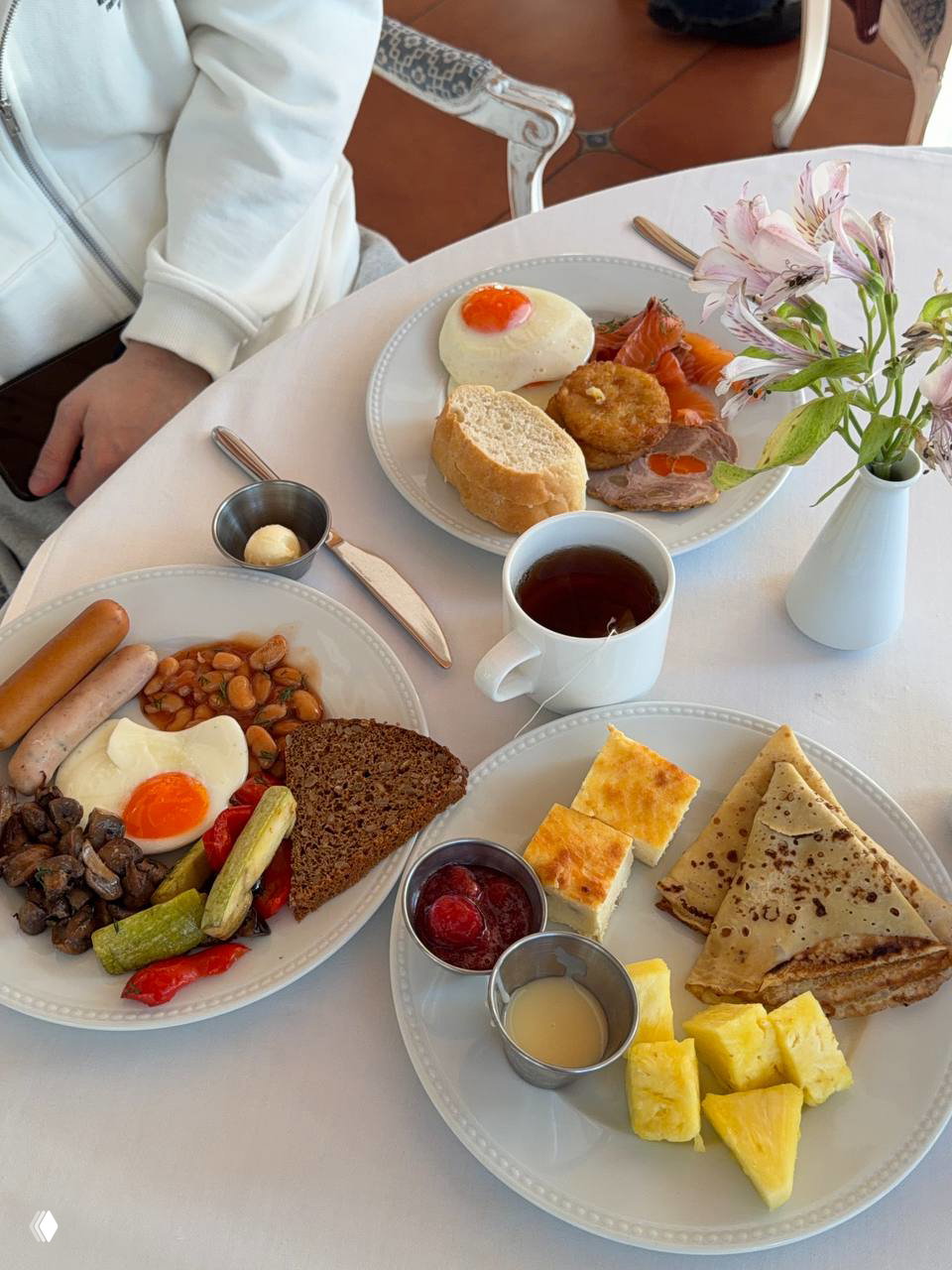 Тарелки со шведского стола: яйца, сосиски, грибы, блины, кусочки ананаса и чашка чая на белой скатерти — завтрак, включённый в проживание в «Скандинавии».