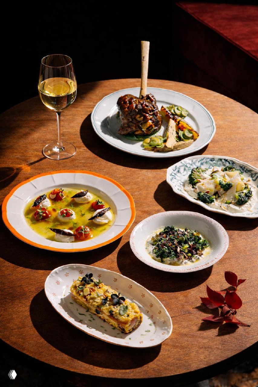 Круглый деревянный стол с несколькими тарелками и бокалом белого вина: ассортимент новых блюд бистро, сервировка на дереве в тёплом боковом свете.