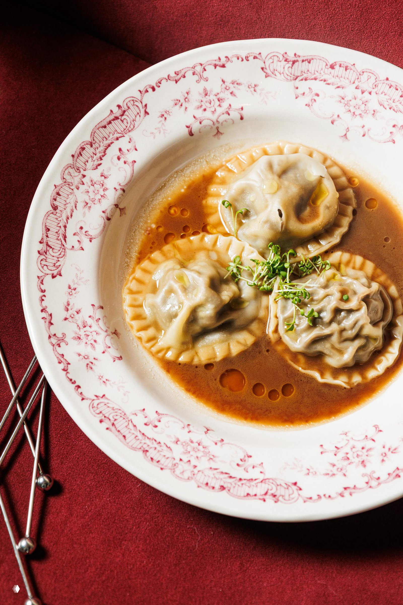 Крупный план равиоли или пельменей в тёмном бульоне в декоративной бело-красной тарелке на бордовом фоне, украшенные микрозеленью.