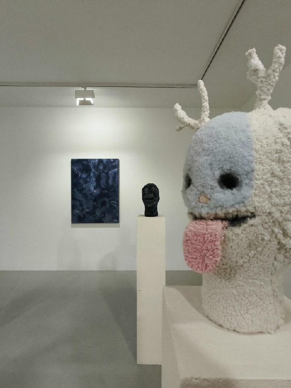 Вид галерейного пространства: на переднем плане пушистая белая скульптура с розовым языком, далее пьедестал и живописное полотно — обстановка экспозиции.