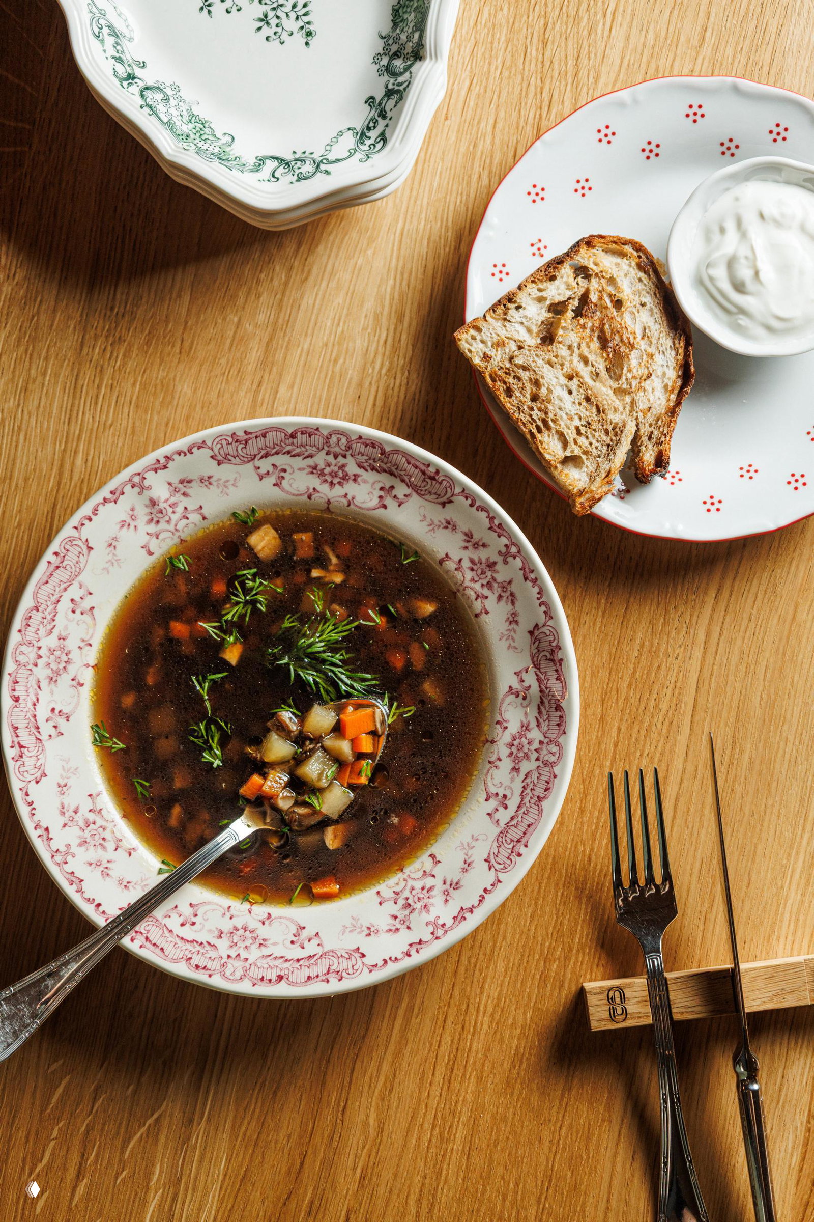 Тарелка с прозрачным супом и мелко нарезанными овощами на деревянном столе рядом с кусочком хлеба и мисочкой сметаны — уютная подача сезонного супа.