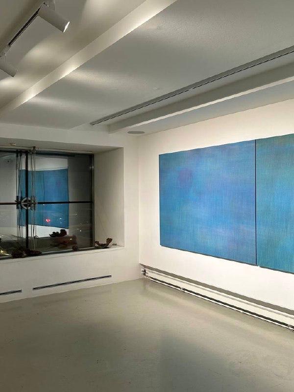 Зал галереи с крупным синим полотном в два холста на белой стене, холодный выставочный свет и вид на окно и низкий радиатор под стеной.