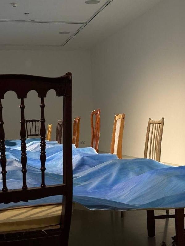 Инсталляция с длинным столом, накрытым полупрозрачной синей тканью, вокруг старинные деревянные стулья — часть экспозиции в галерее.