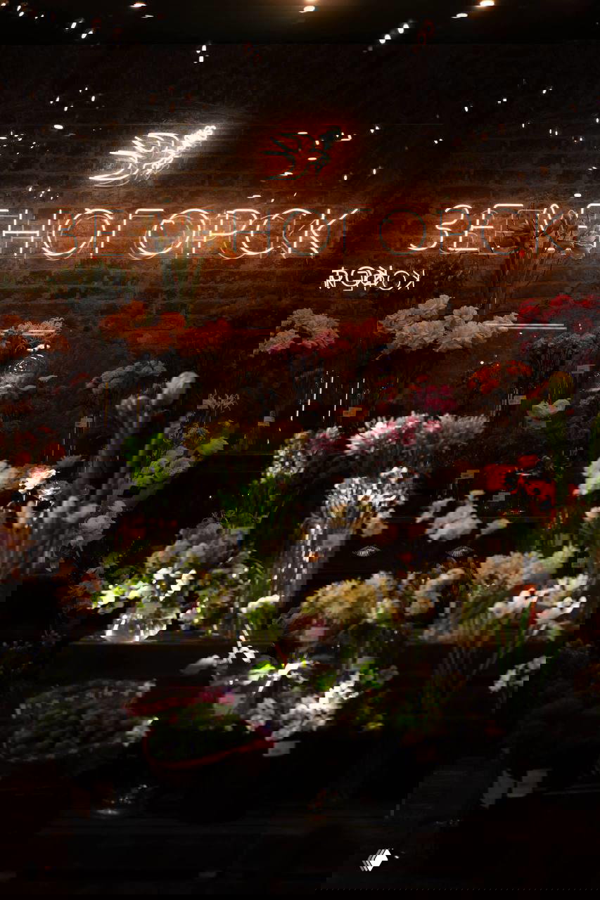 Интерьер цветочной лавки с неоновой вывеской «Зеленогорск» и множеством букетов и ваз на прилавке в тёплой витрине.