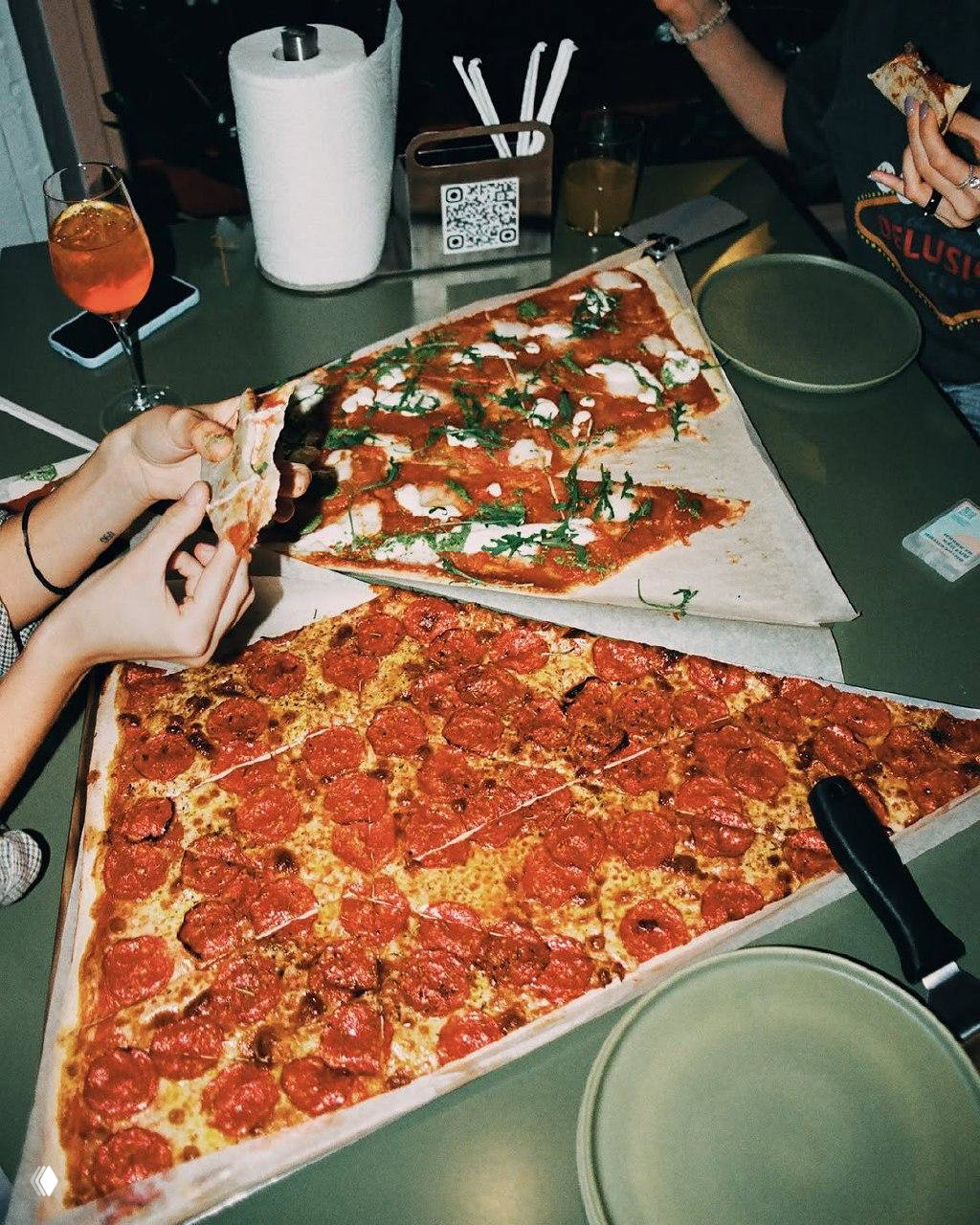Две огромные треугольные пиццы на зелёном столе в кафе Oversize Pizza Club, руки тянут ломтики, рядом тарелки, стаканы, держатель салфеток и QR‑код на подставке.