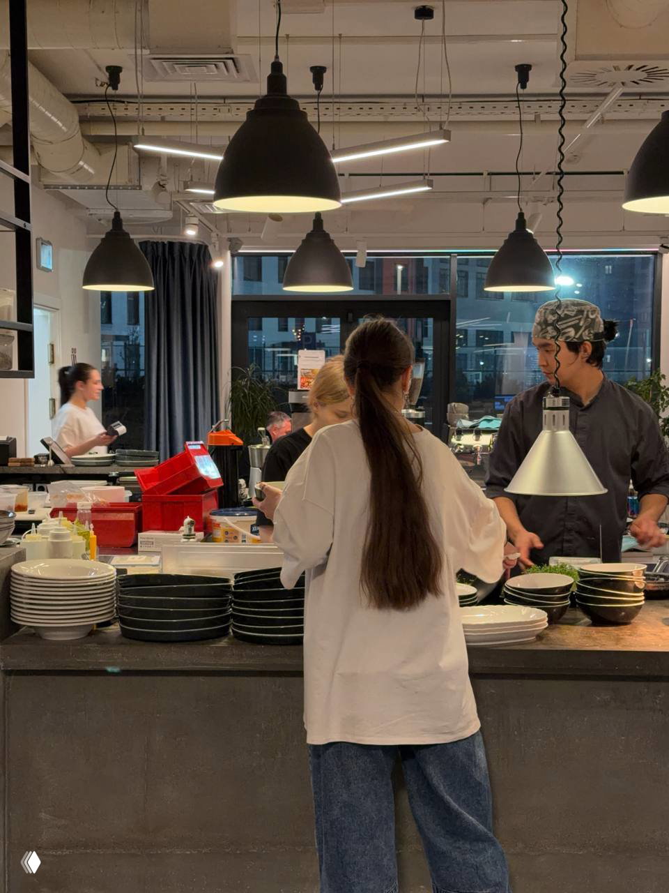 Кухонная зона с поварами и стойкой в бистро Pajama: открытая кухня, подвесные светильники и вид на процесс приготовления блюд.