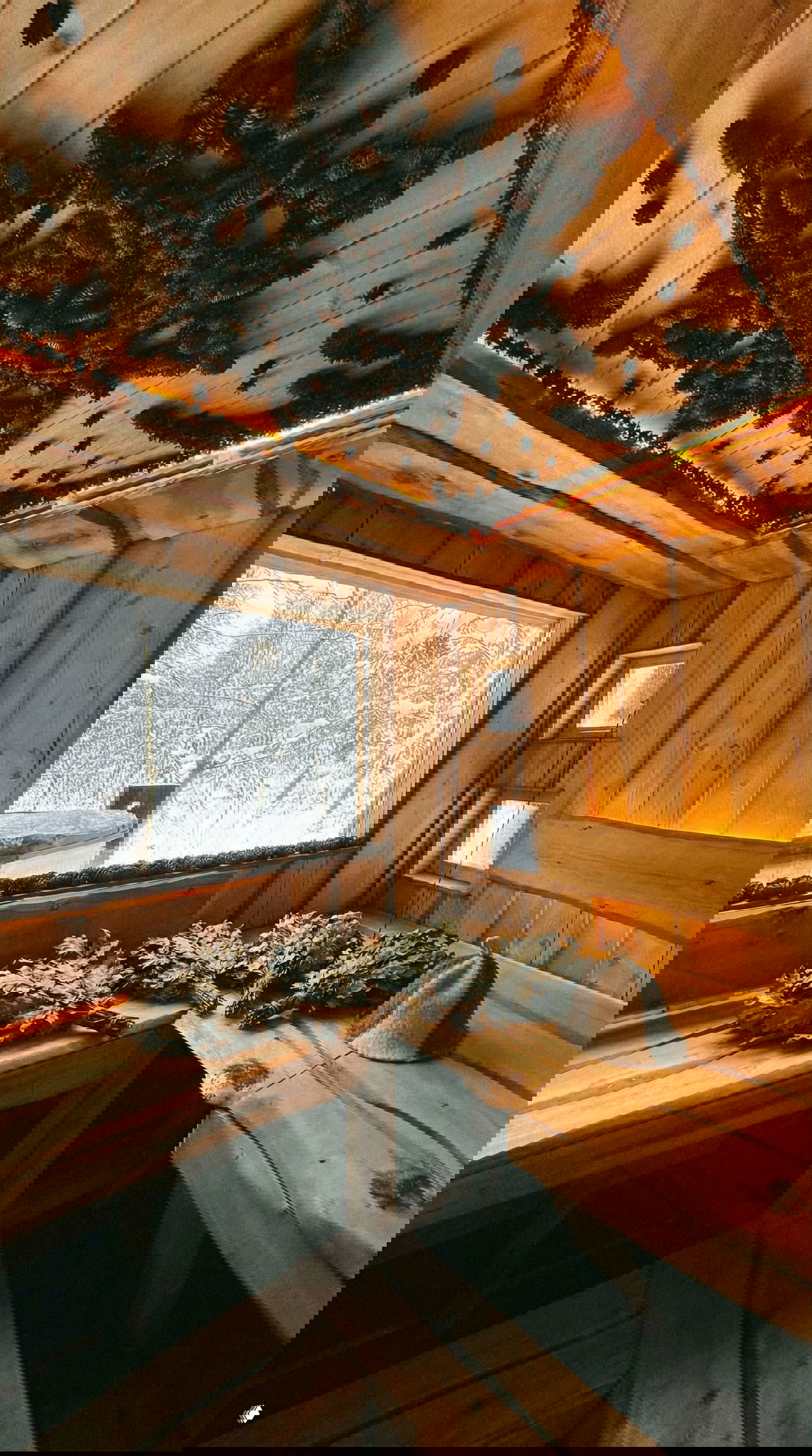 Деревянная парная в Lembo: лавки, связки веников и традиционная шапка для бани на фоне окна с видом на заснеженный лес и купель снаружи.