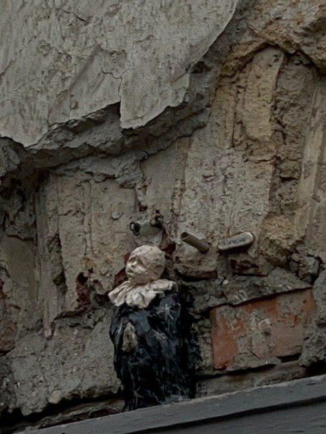 Крошечная скульптура в образе буффона или клоуна в тёмно-белом костюме, вписанная в облупившийся участок стены под карнизом здания; Санкт-Петербург, автор — Екатерина Березина.