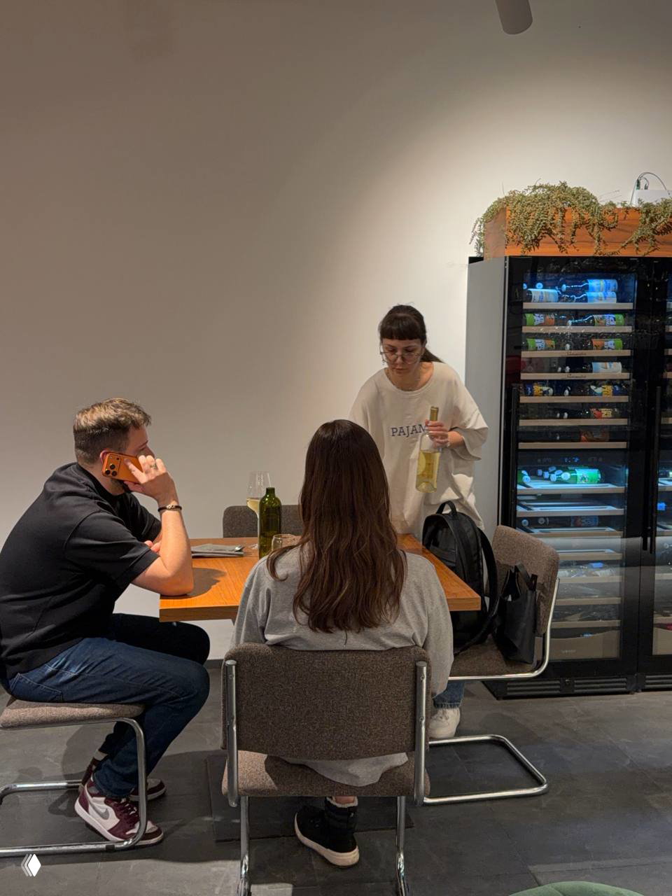 Официантка в футболке с логотипом Pajama подаёт бутылку вина за столом, видны холодильник для вин и спокойный минималистичный интерьер.