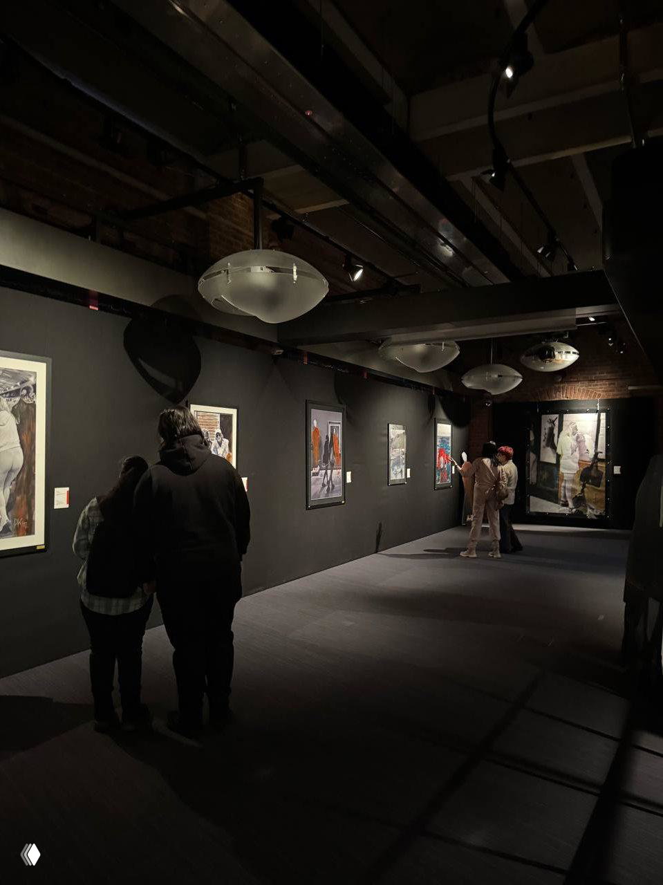 Тёмный зал экспозиции с полотнами на чёрных стенах, группы посетителей у работ и направленное освещение, создающее камерную атмосферу.