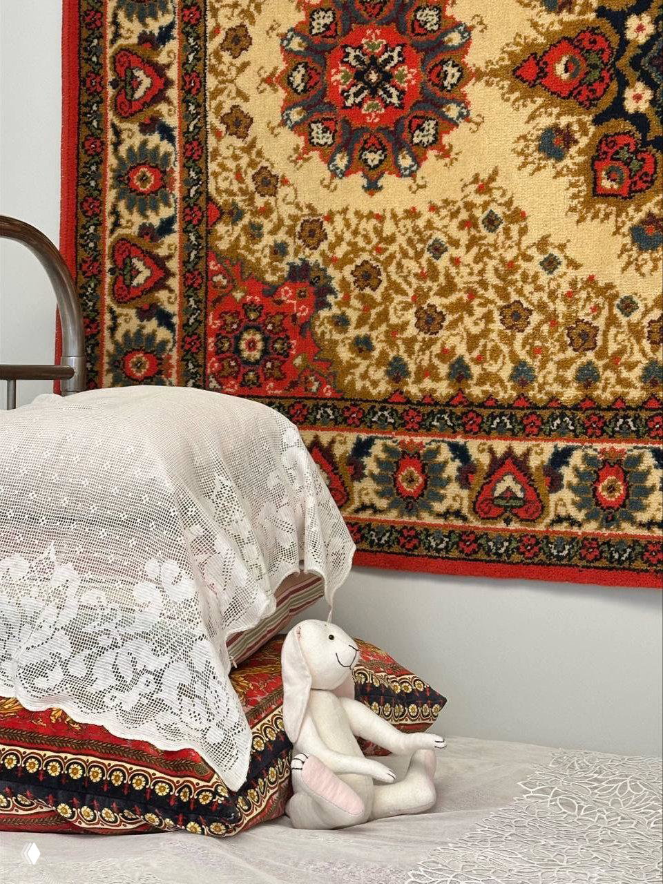 Инсталляция домашней сцены: ковёр на стене, кружевная покрывка на подушках и мягкая игрушка на кровати — образ семейной памяти в экспозиции.