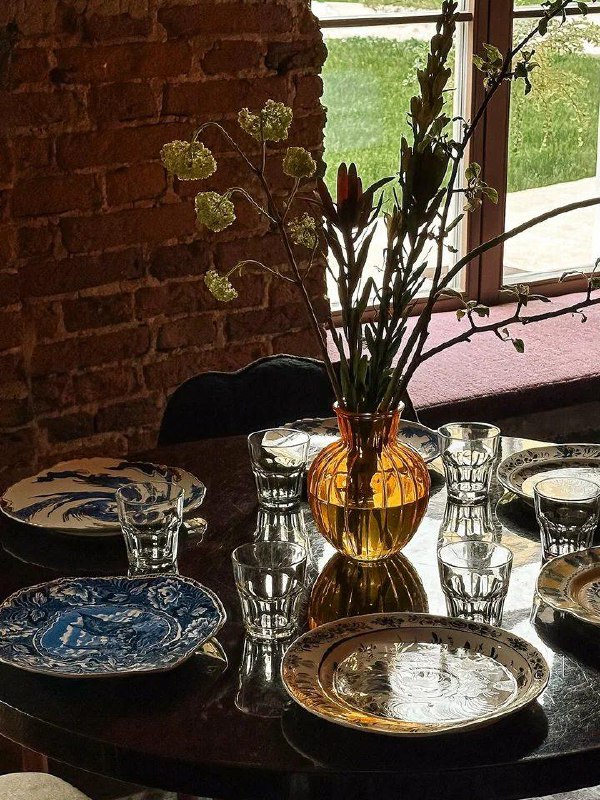 Сервировка столика в Jam Cafe: фарфоровые тарелки, стеклянные стопки и ваза с цветами на фоне кирпичной стены у окна.