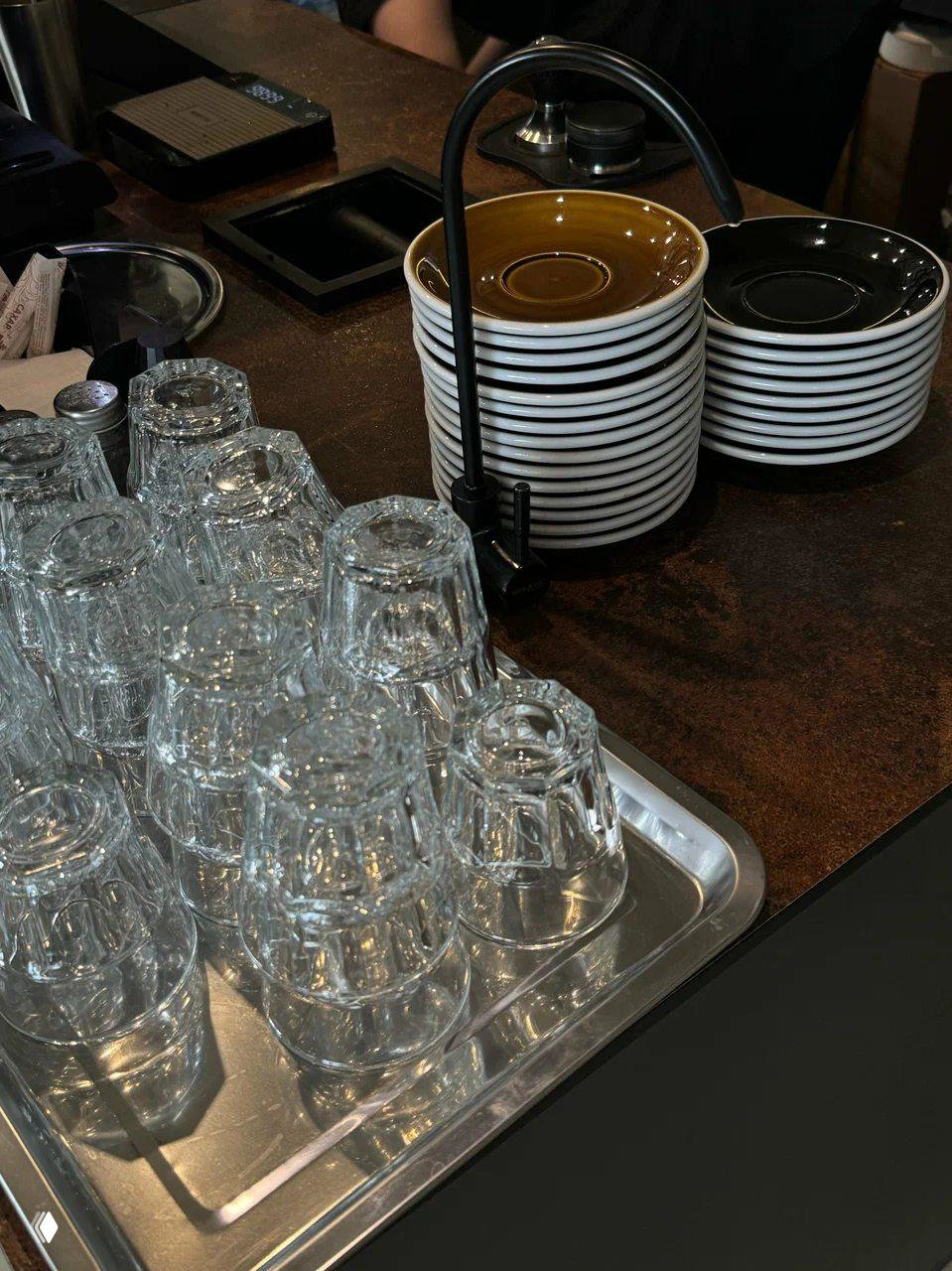 Стеклянные стаканчики и стопка чайных блюдец на барной стойке кофейни Keny — вид из зоны выдачи напитков, акцент на посуде.