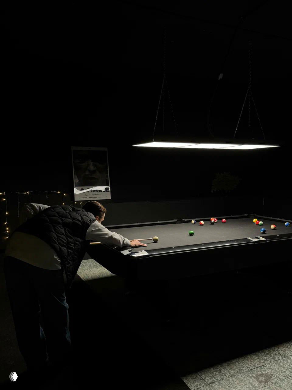 Игрок наклоняется над чёрным пул-столом в тёмном зале с подвесными светильниками, видно шаровые наборы и камерную атмосферу клуба
