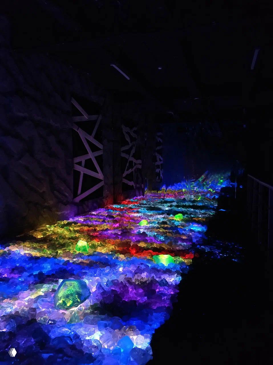 Полоса «кристаллических» камней и гранул, подсвеченная радужно‑неоновыми огнями, образует светящийся путь внутри тёмного зала выставки.