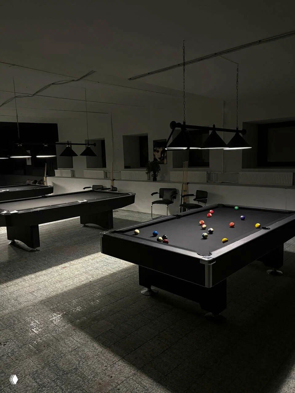 Интерьер бильярдной с несколькими чёрными столами, подвесными лампами и зеркалами на стене — строгий монохромный стиль пространства