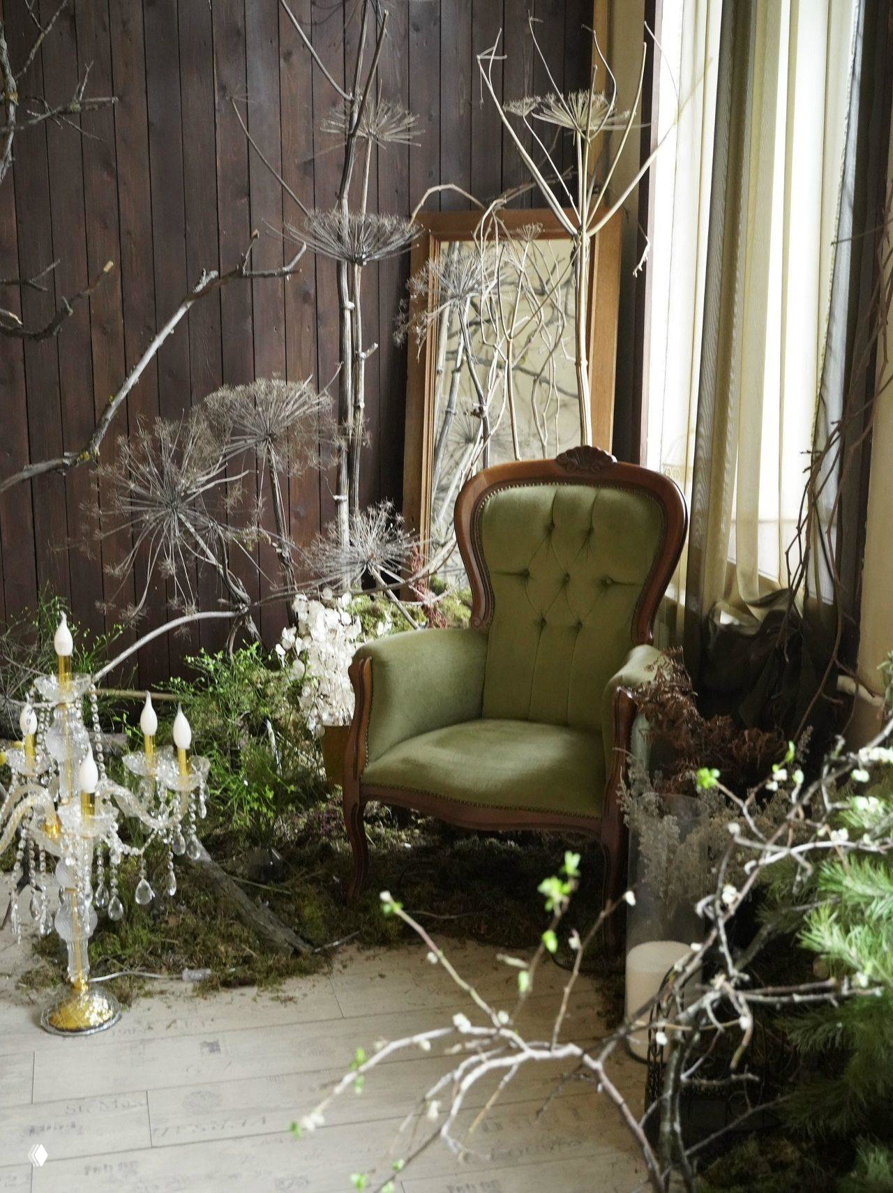 Уютный уголок с зелёным креслом, хрустальной люстрой и сухими ветвями на фоне деревянной стены и мха на полу.