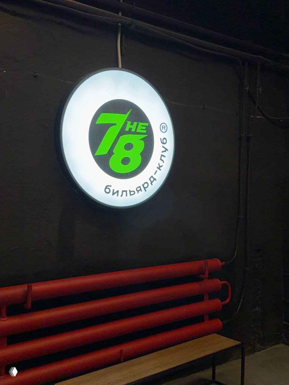 Подсвеченный логотип «7 не 8» на тёмной стене у входа бильярд‑клуба — круглый неоновый знак и индустриальный декор рядом.
