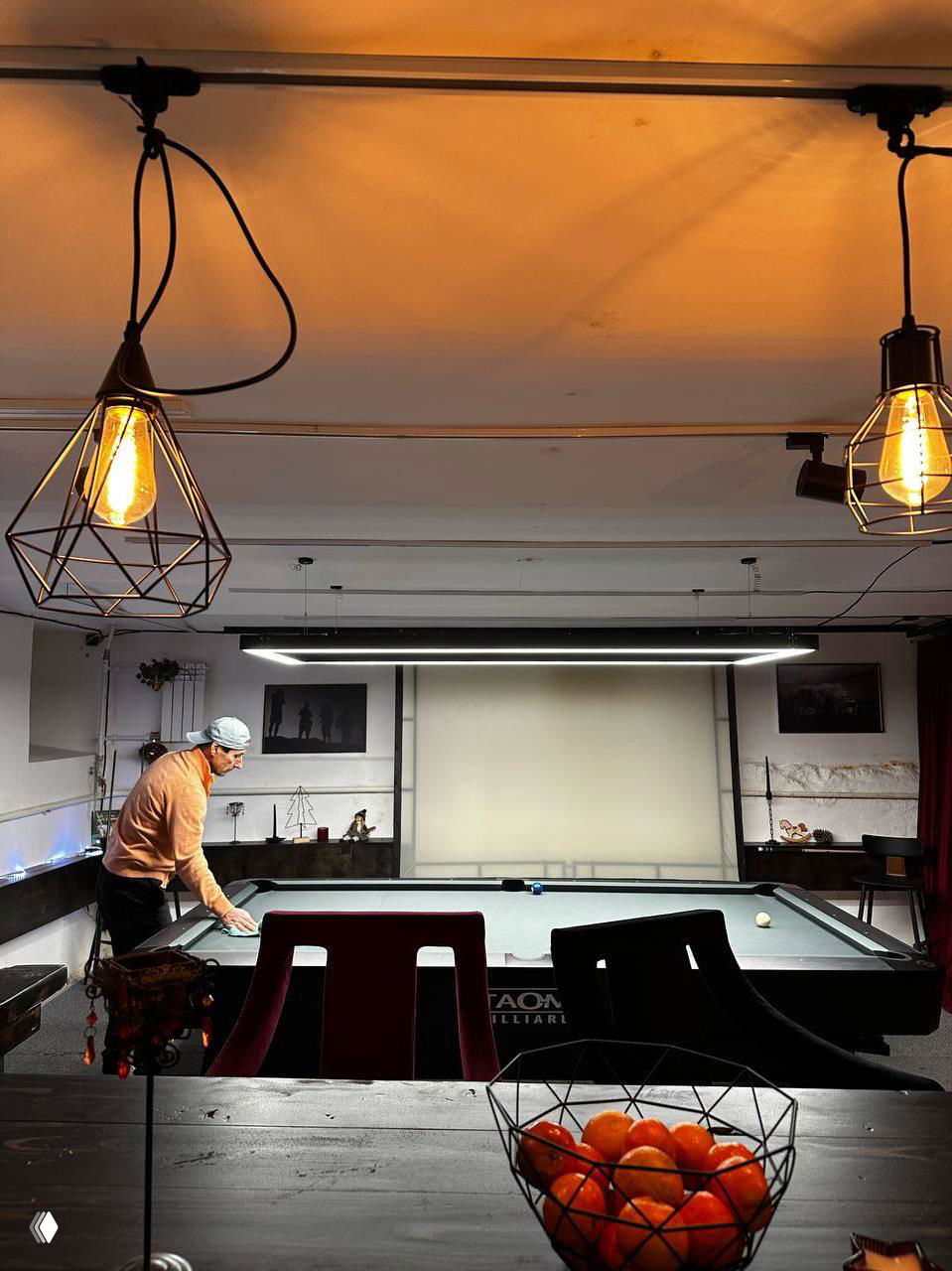 Человек готовится к игре за бильярдным столом, в переднем плане миска с мандаринами и лампы — тёплая сцена бара в клубе перед партией