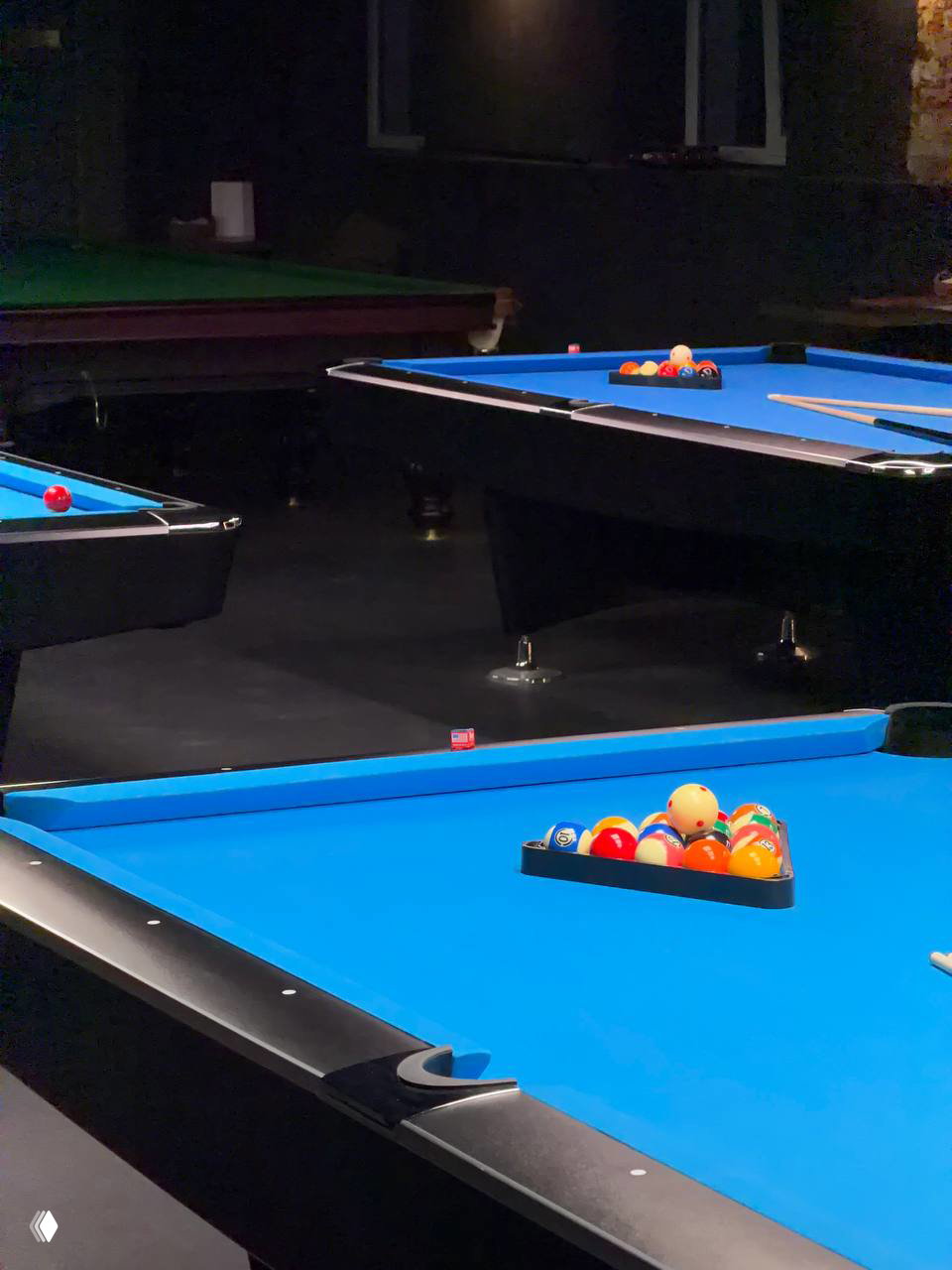 Несколько бильярдных столов с синим сукном в затемнённом зале, на столах расставлены шары и кии — общий план игровой зоны клуба.