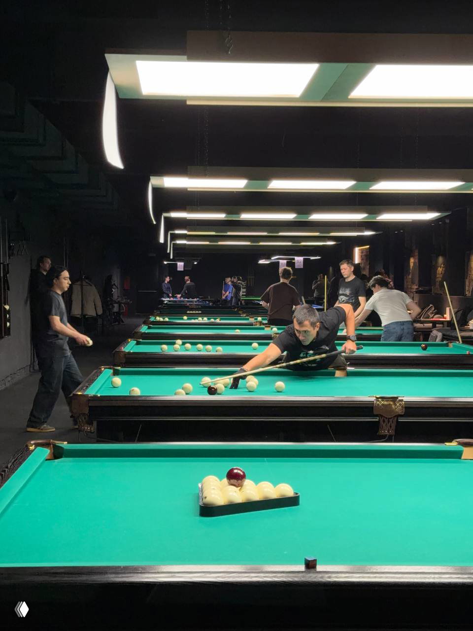 Ряд синих бильярдных столов в клубе, на переднем плане треугольник с шарами, фон — просторный и затемнённый зал для игры.