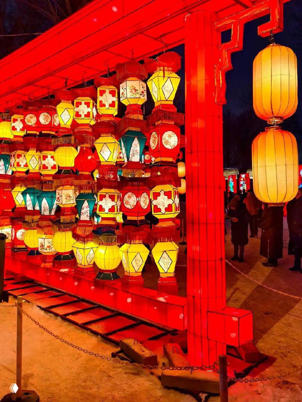 Стена из разнообразных подвесных разноцветных фонарей в красной деревянной раме, аллея фестиваля с прохожими и огнями.