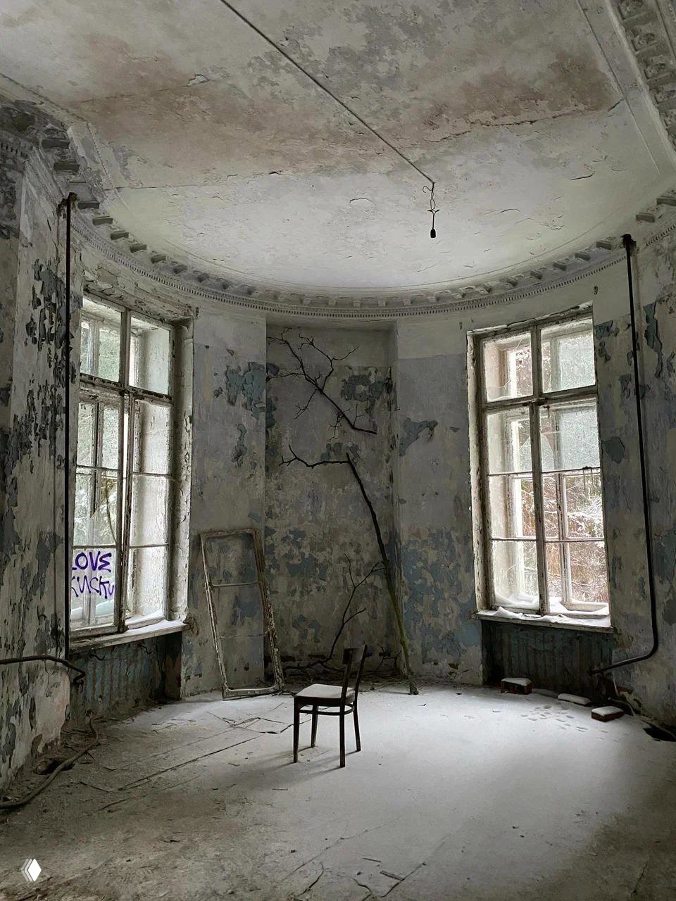 Круглая комната заброшенной усадьбы с облупившимися стенами, пыльным полом, одиноким стулом и старой рамой у окна.
