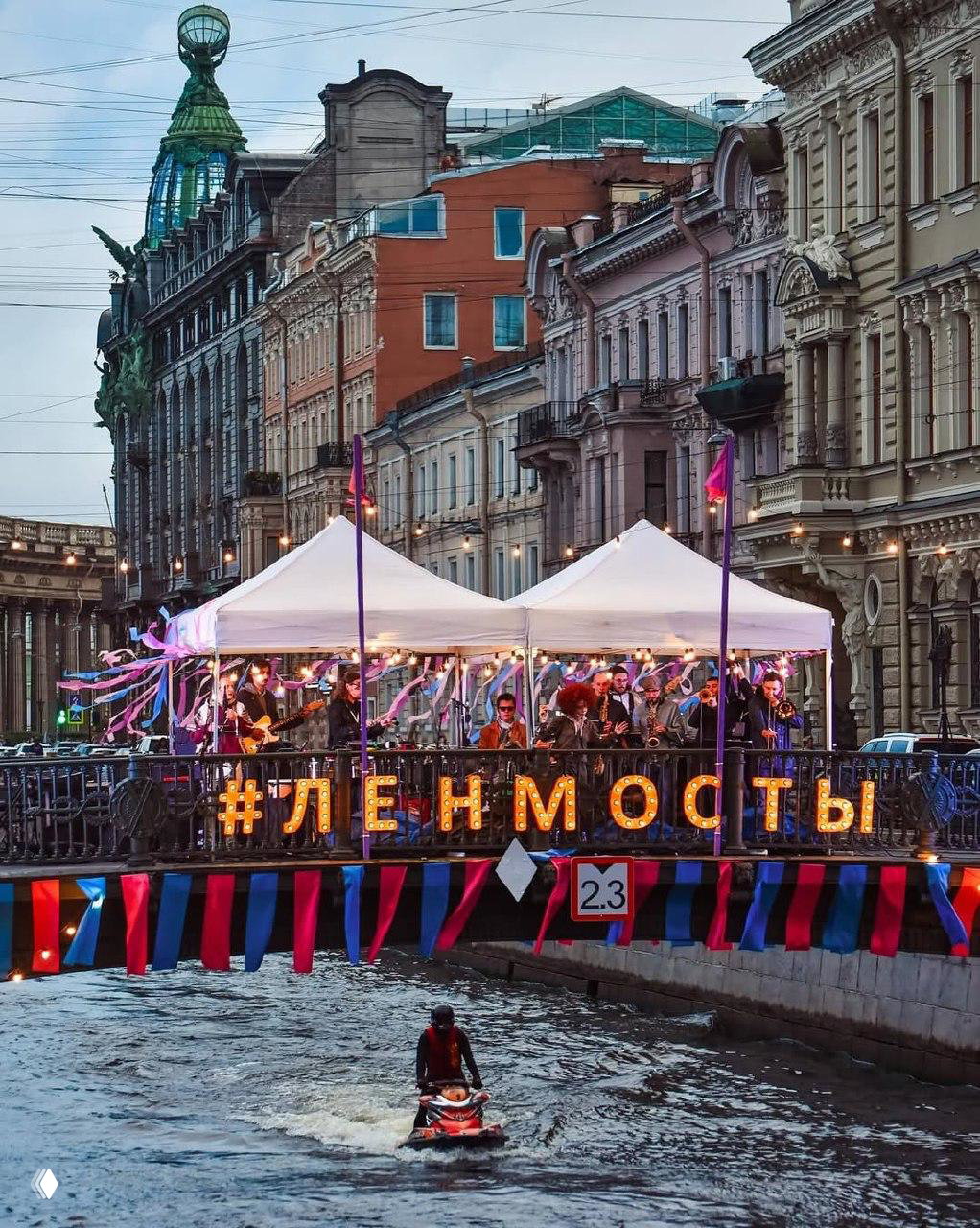 Широкий план моста с большой светящейся инсталляцией «#ЛЕНМОСТЫ», сценой и зрителями вдоль набережной в городской атмосфере.