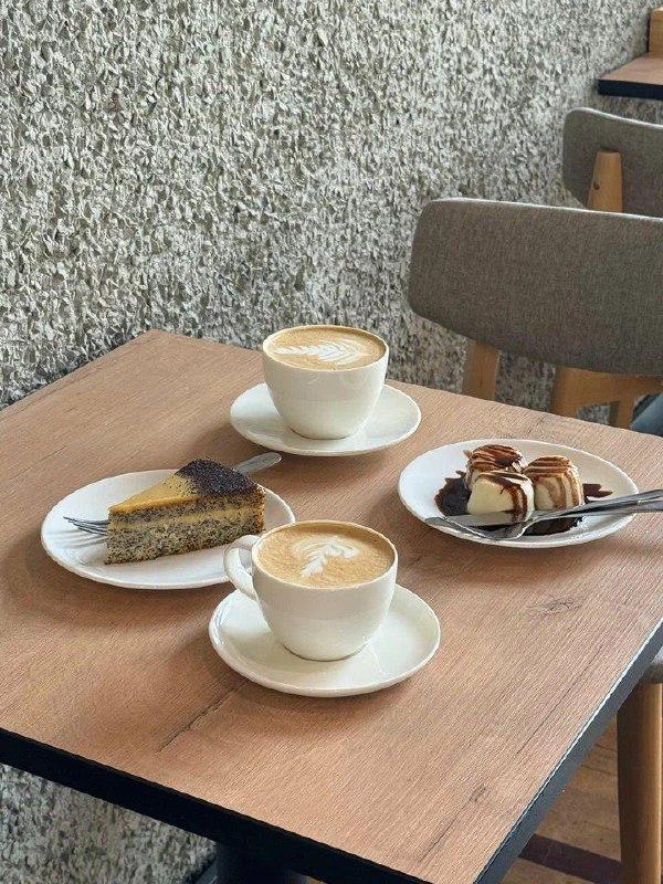 На столе в кофейне две чашки капучино, кусок пирога и тарелка с десертом на деревянной поверхности — типичный кофейный сет.