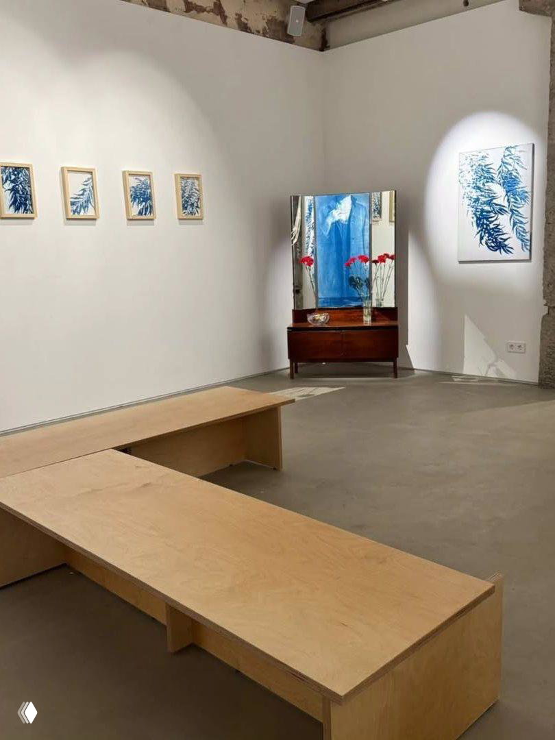 Широкий кадр экспозиции: деревянные скамьи, белые стены с серией небольших синих работ и угол с зеркалом и комодом.