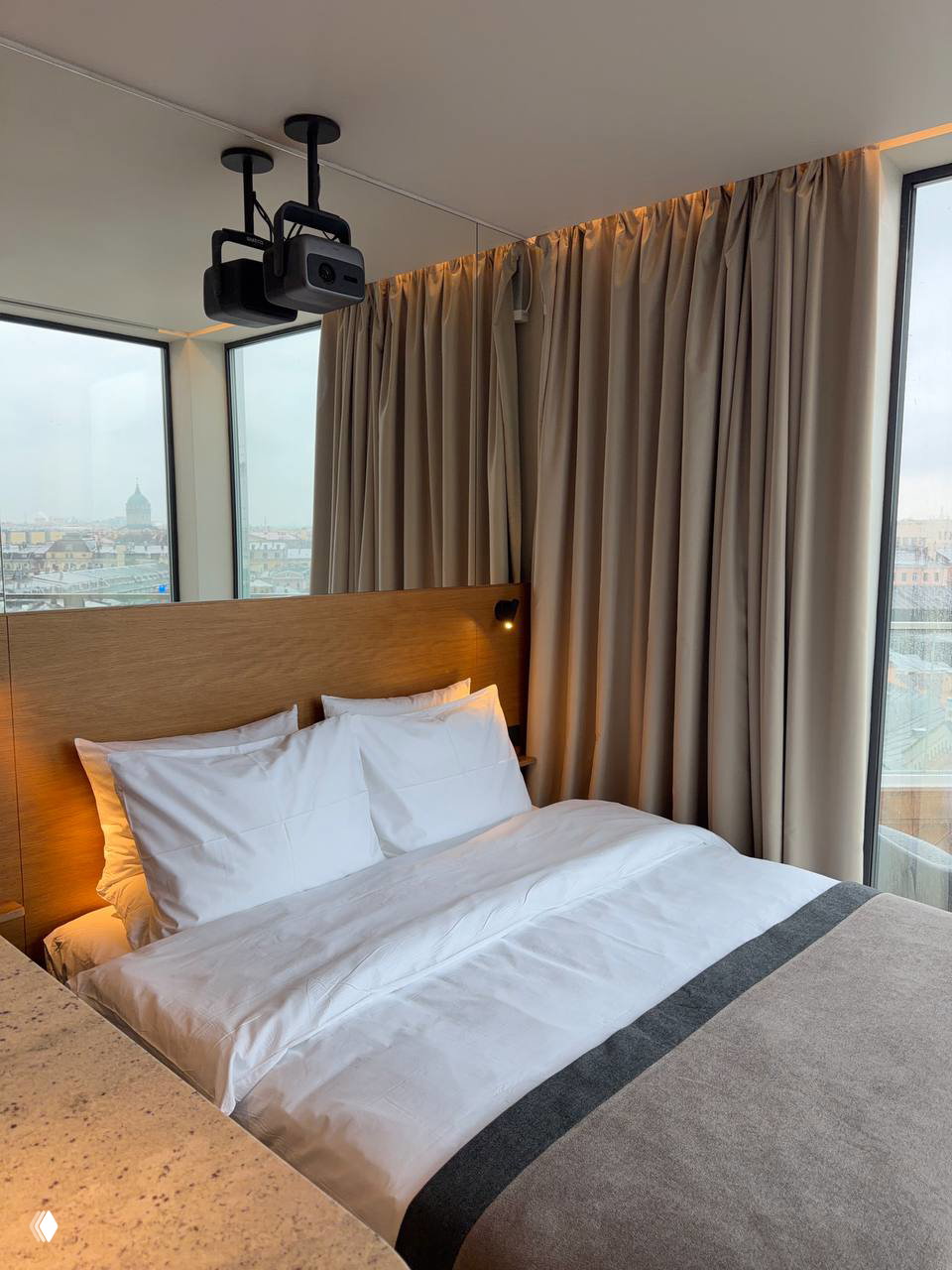 Угол спальни у панорамного окна: кровать рядом с большим окном, мягкие текстуры, шторы и вид на город — минималистичный дизайн.