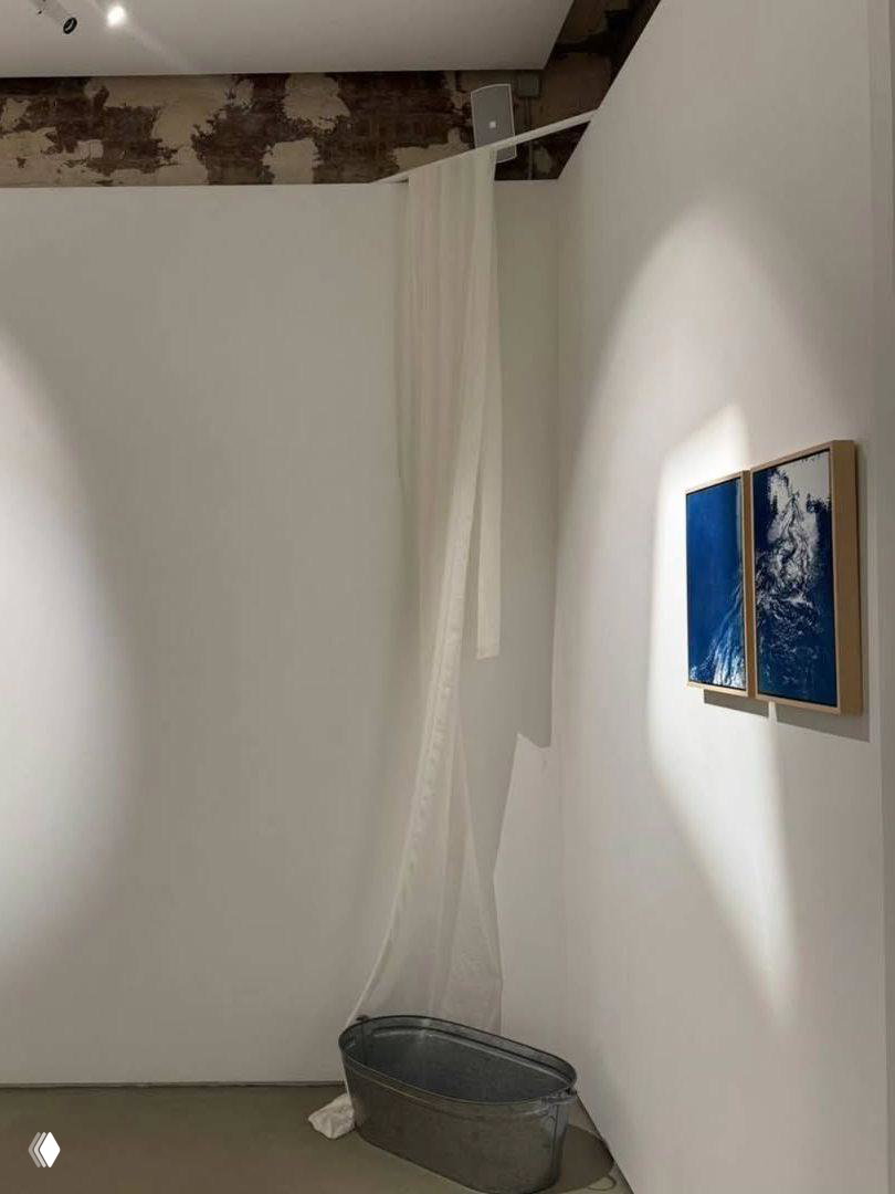 Угол зала Open You Gallery: подвешенная прозрачная ткань, металлическая ванна на полу и пара маленьких синих работ на стене.