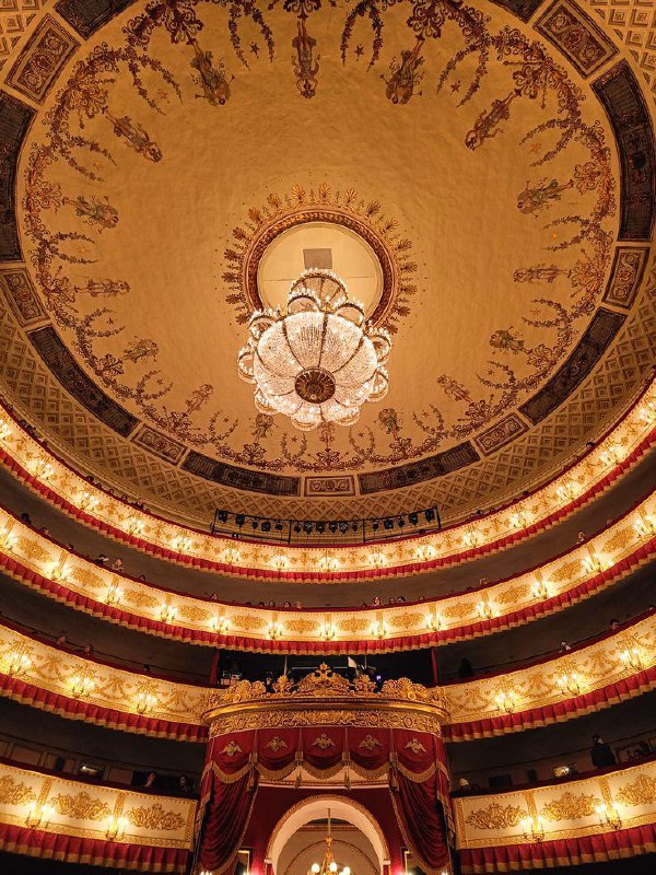 Вид вверх на купол и хрустальную люстру исторического театра, окружённый ярусами и освещёнными балконами с орнаментом.