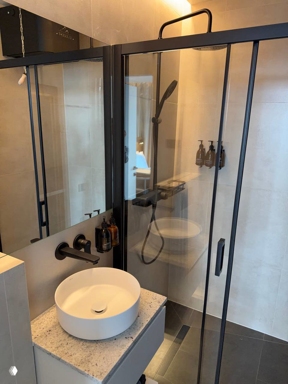 Ванная комната в глэмпинге: раковина на столешнице, большое зеркало и душ с чёрной рамой, аккуратно размещённые флаконы с косметикой.