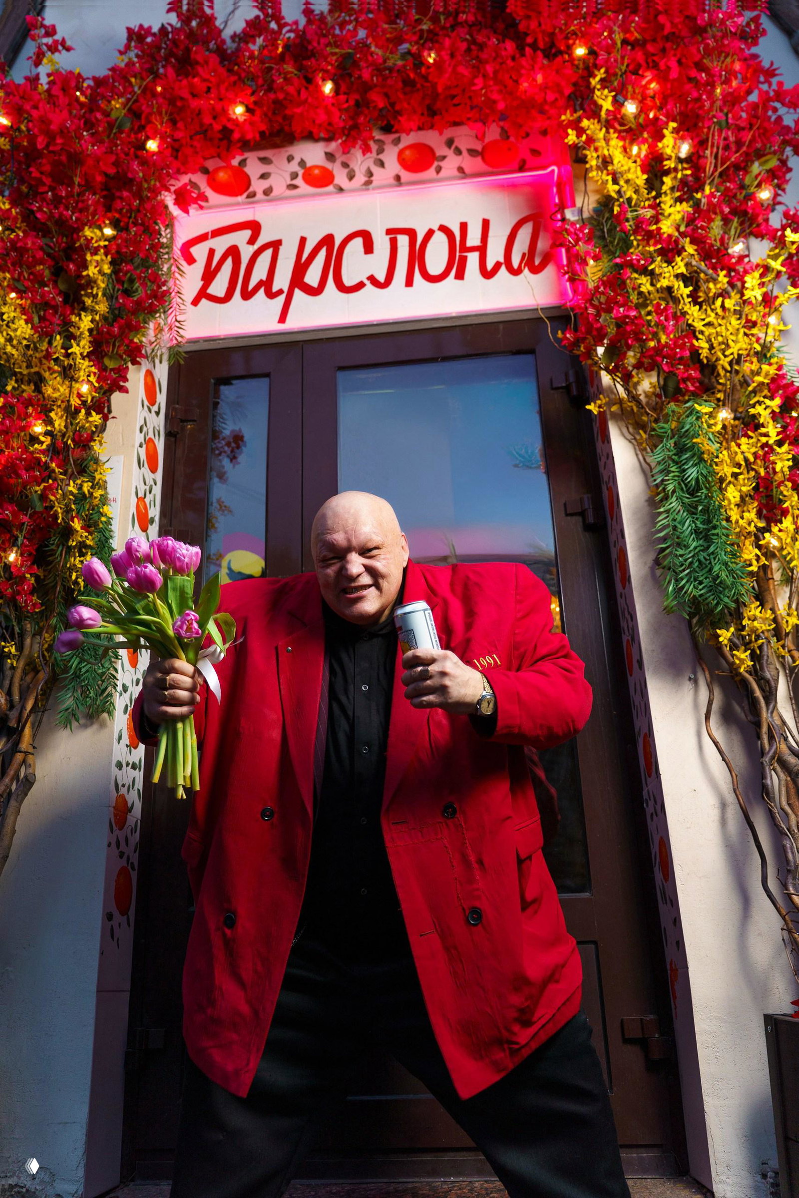 Мужчина в красном пиджаке у входа в ресторан держит букет тюльпанов и банку напитка; вокруг праздничная флористика и вывеска заведения.