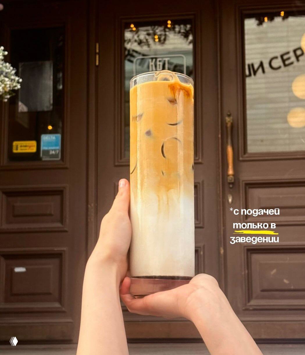 Фотография литрового латте/бамбла, удерживаемого двумя руками на фоне двери кофейни КБТ; виден яркий молочно-кофейный градиент.