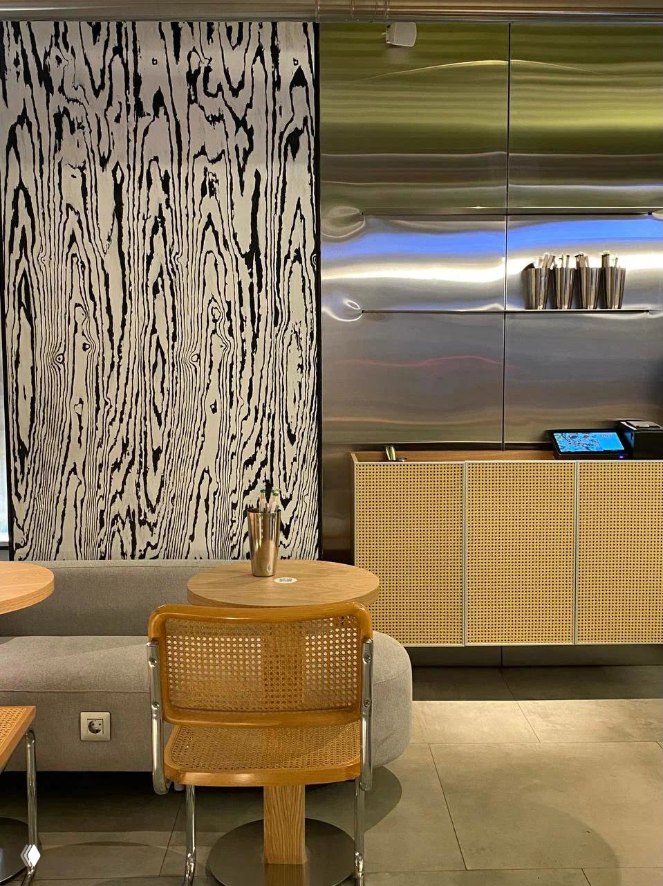 Интерьер кафе Kuta: минималистичная зона с плетёными креслами, столиками и стойкой на фоне металлической панели и декоративного рисунка