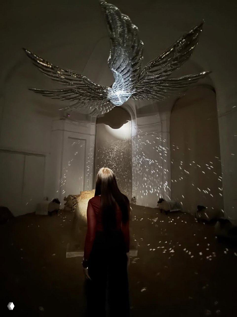 Инсталляция с подвесной фигурой ангела: зеркальные перья и световые блики в затемнённом пространстве, посетитель наблюдает инсталляцию.