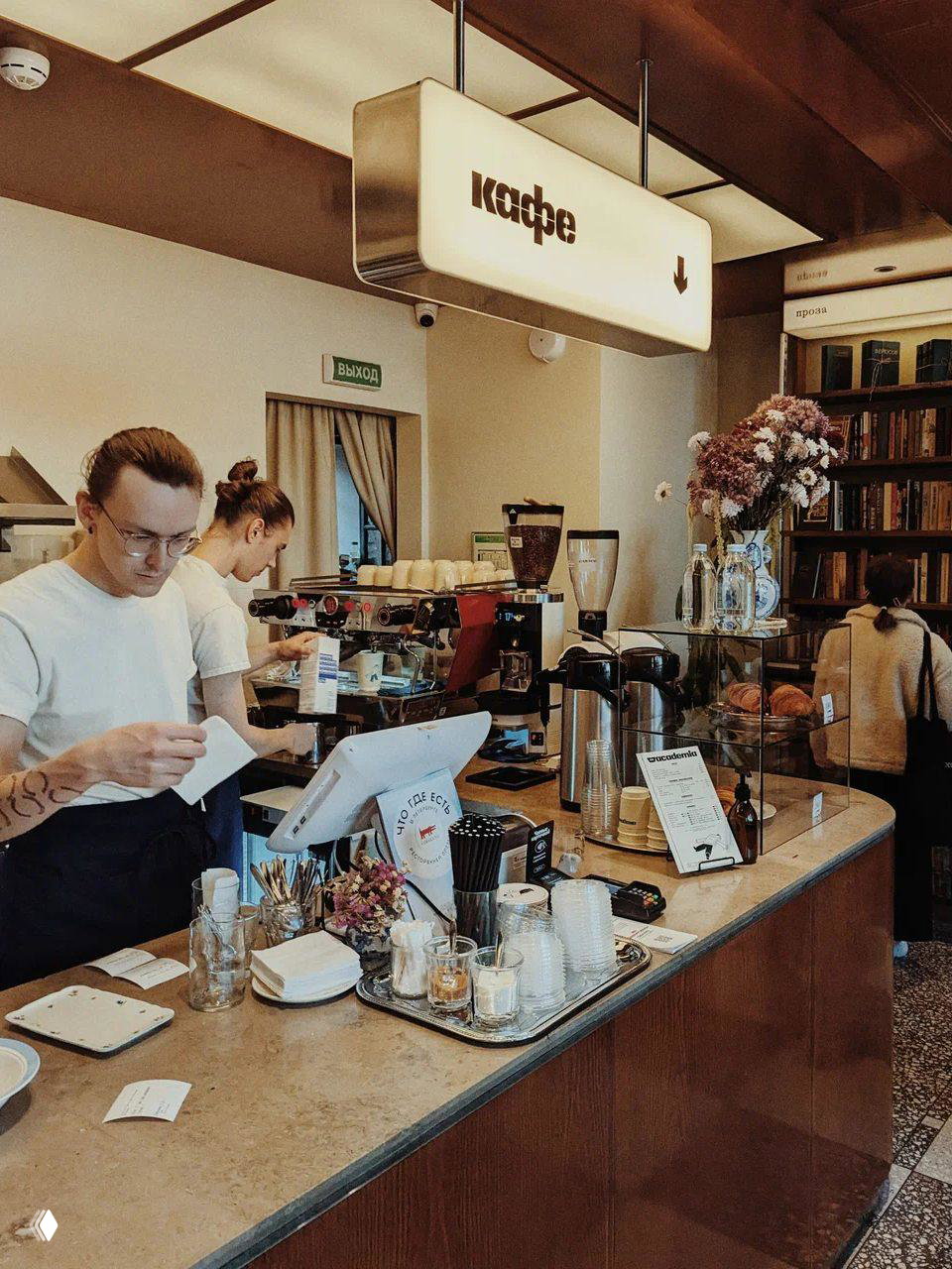 Кофейная стойка и бариста в Tagliatella Cafe внутри книжного пространства: чашки, кофемашина и витрина с выпечкой на заднем плане.