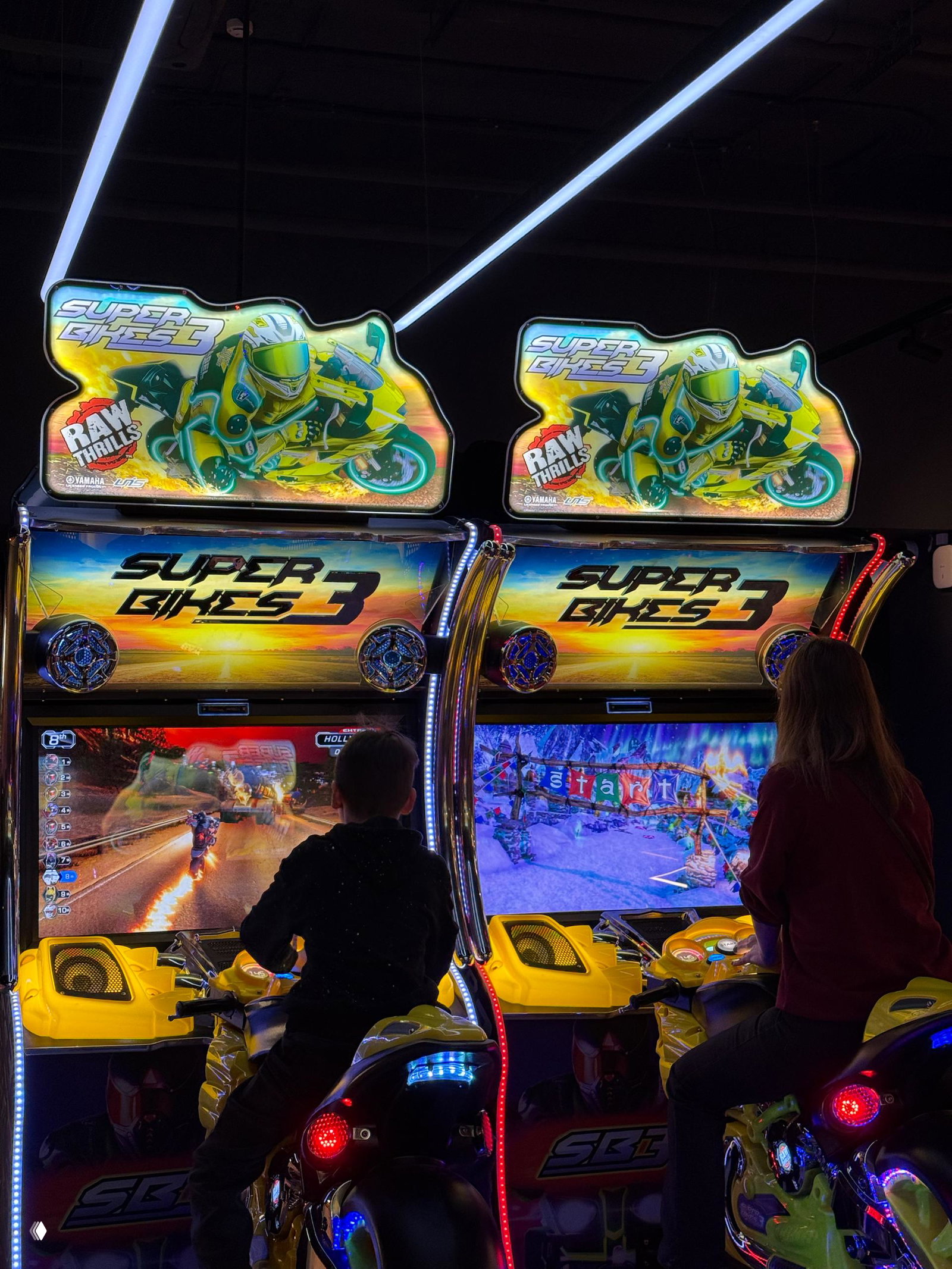 Дети и подростки за игровыми мото-симуляторами и гоночными автоматами: неоновая вывеска и экраны создают атмосферу аркадных гонок.