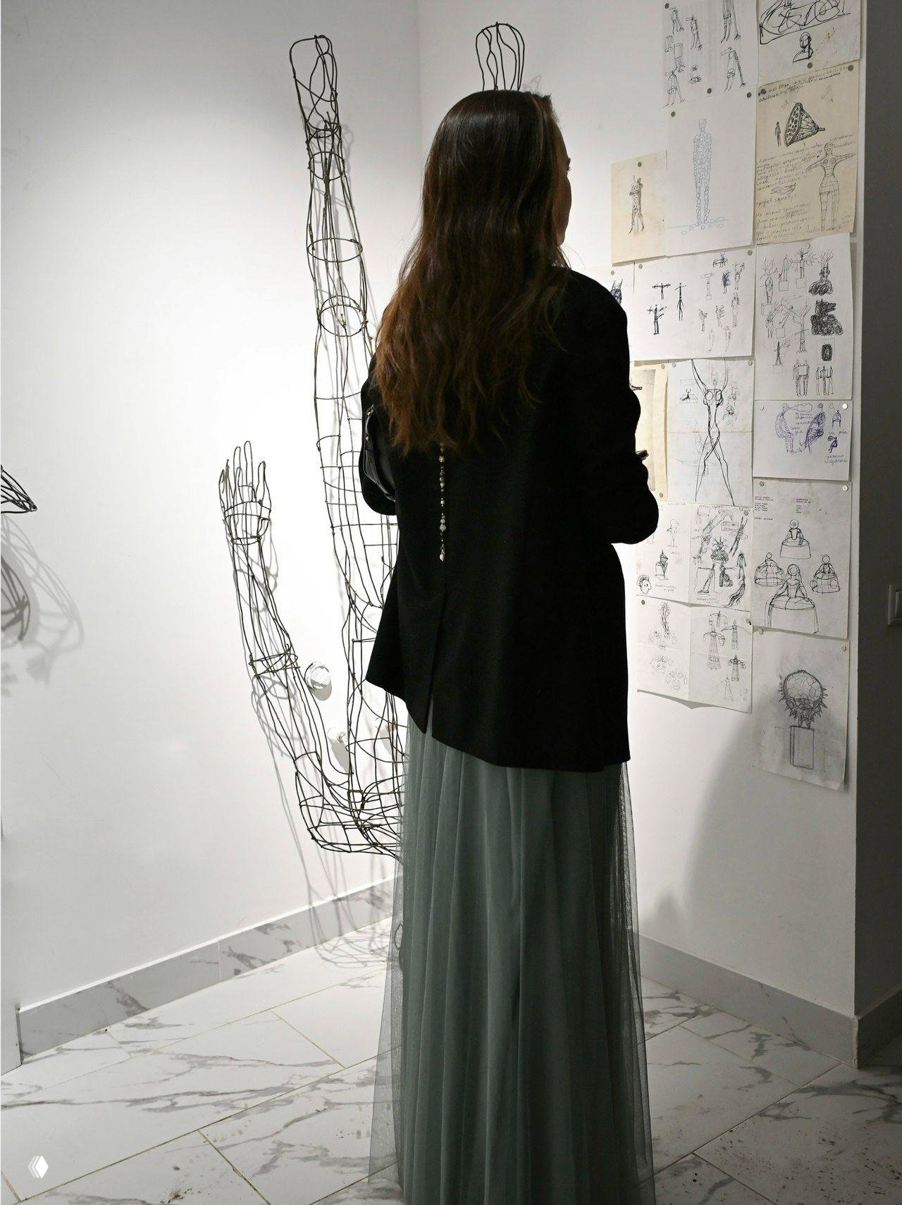 Женщина‑посетительница стоит спиной, рассматривая эскизы и проволочную инсталляцию; вид на композицию и тени на полу.