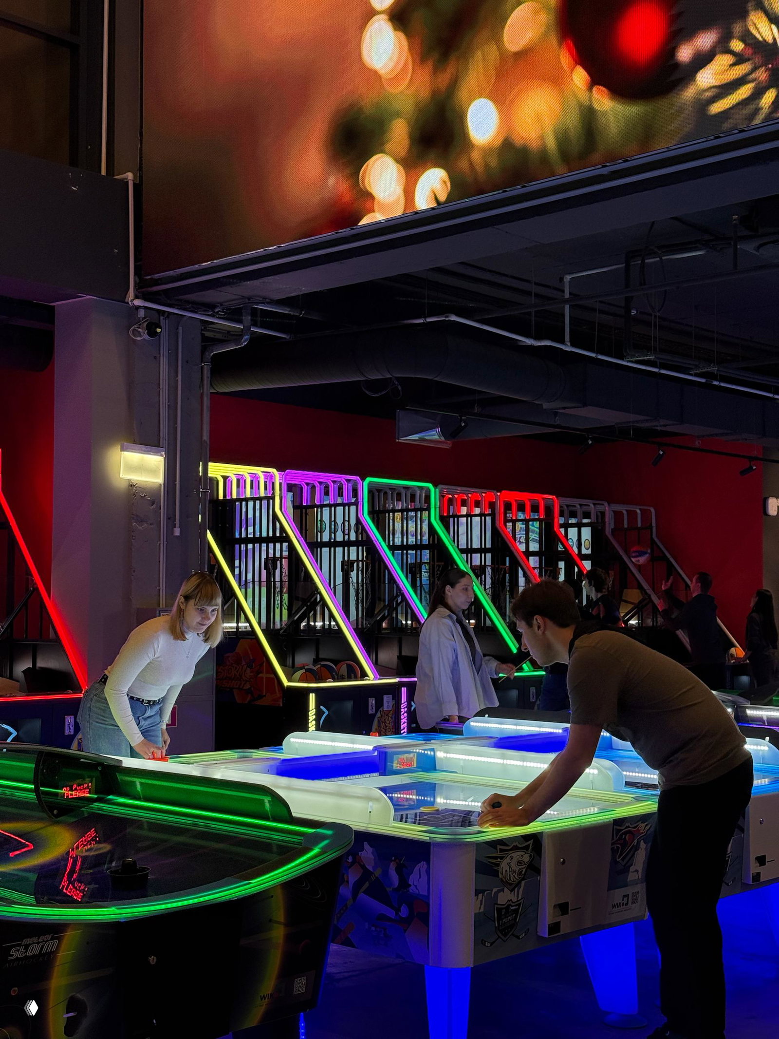 Столы для аэрохоккея и другие настольные аркады с неоновой подсветкой: люди играют в тематическом развлечении в интерьере MazaPark.