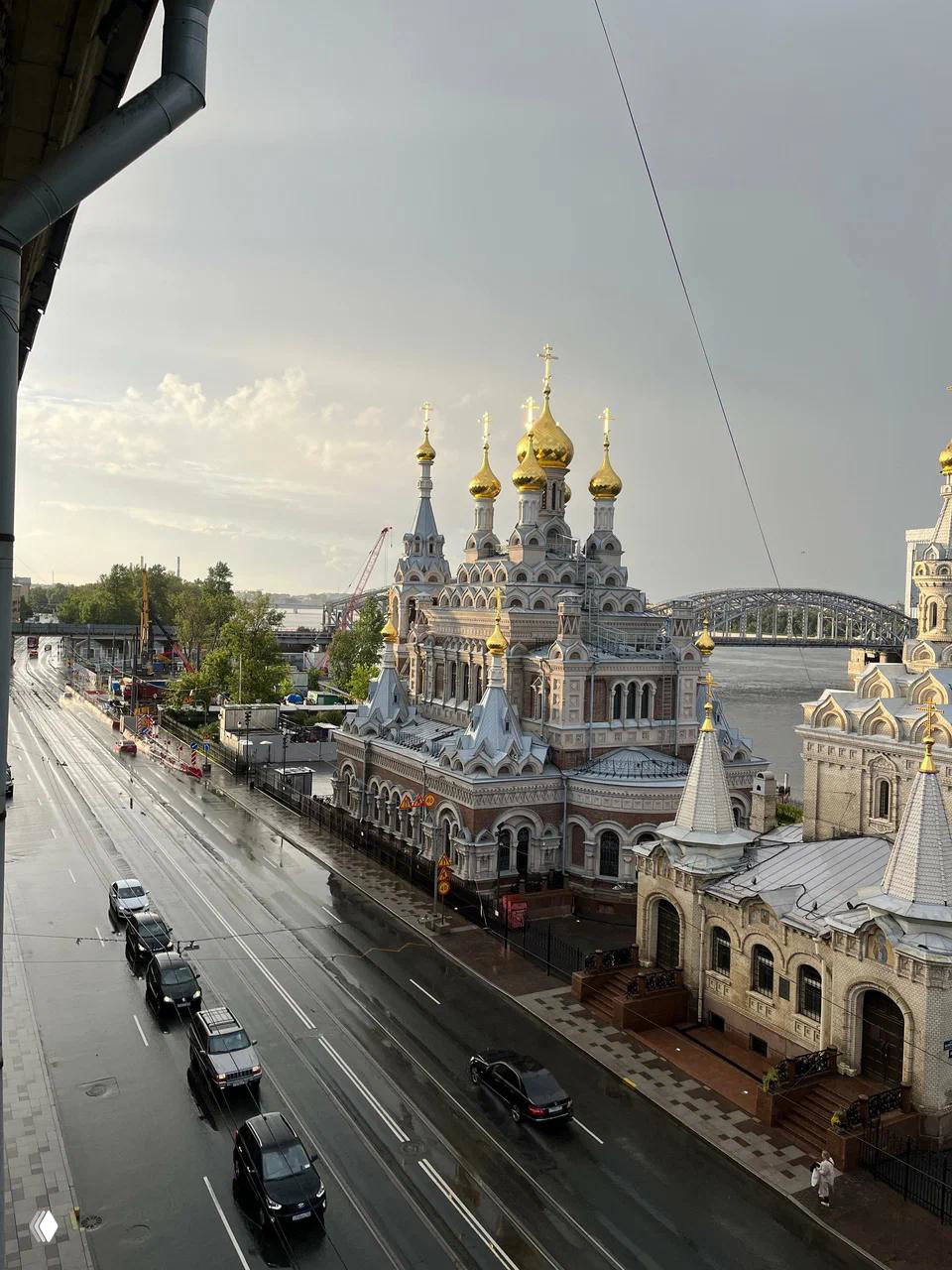 Вид на Скорбященскую церковь с улицы после дождя: дорога, здания, Нева и мост на фоне облачного неба, городской контекст.
