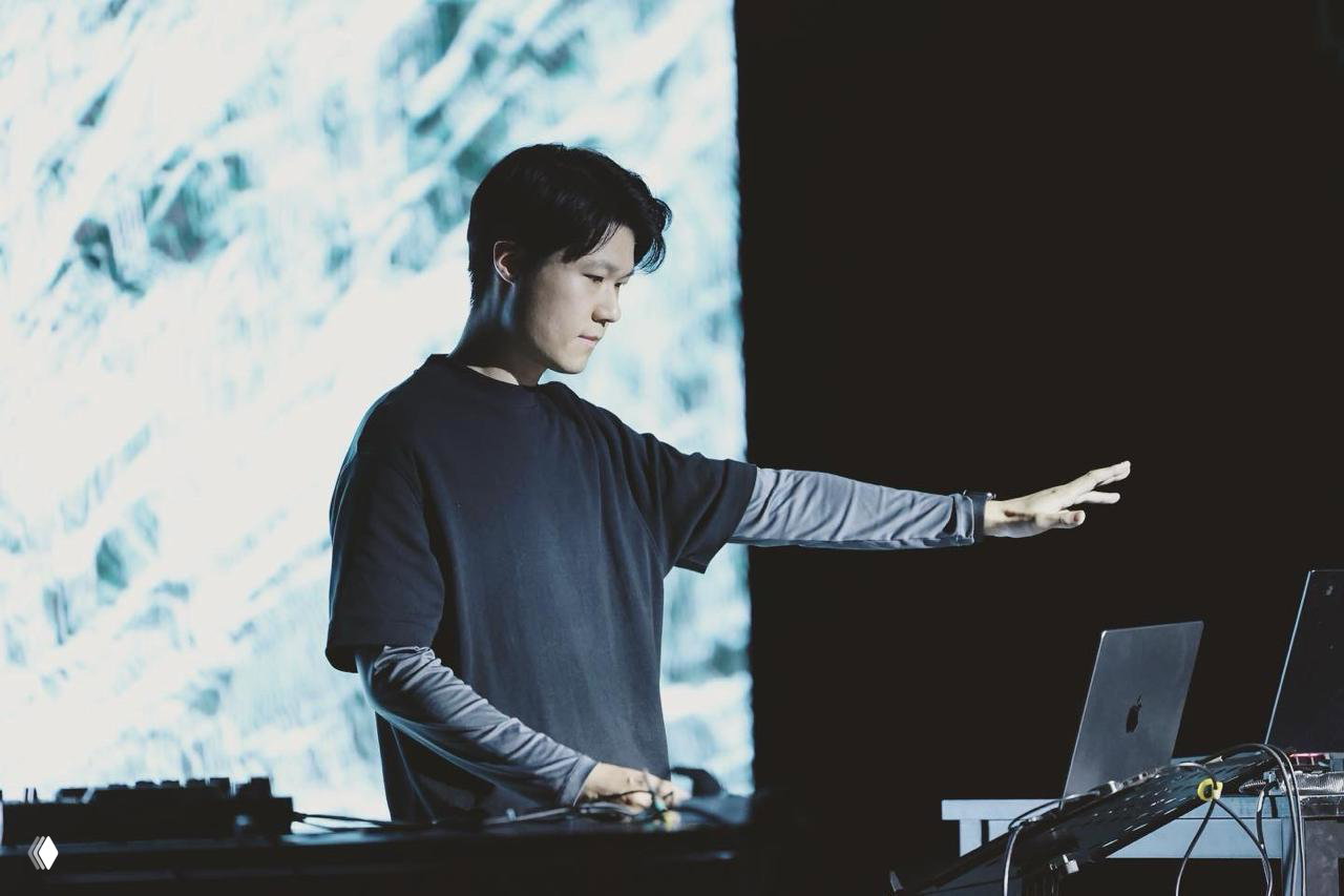 Медиахудожник Шихуа Ма во время выступления за ноутбуком и контроллерами на сцене, акцент на жесте и световой проекции позади исполнителя.