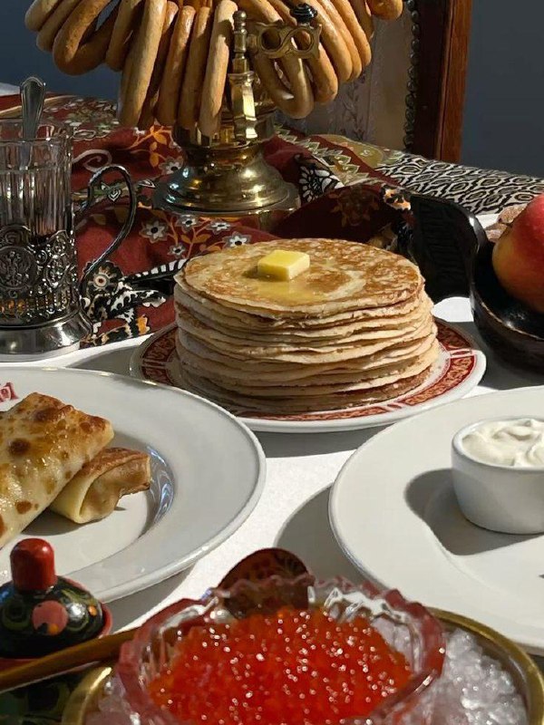 Праздничный стол с традиционными угощениями: стопка тонких блинов с маслом, рулетики, красная икра и сметана на тарелках.