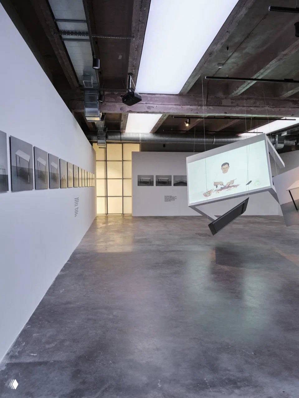 Общий вид экспозиции в галерее: просторный белый зал, висящие объёмные конструкции-экраны и серия фотографий вдоль стены.