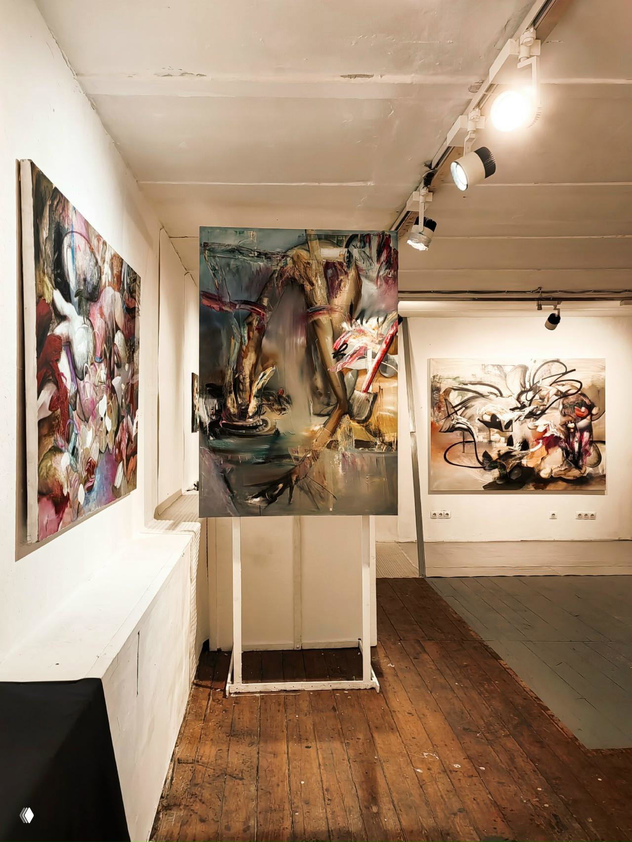 Общий вид галерейного пространства с деревянным полом, несколькими картинами и центральной вертикальной инсталляцией, создающей зонирование экспозиции.