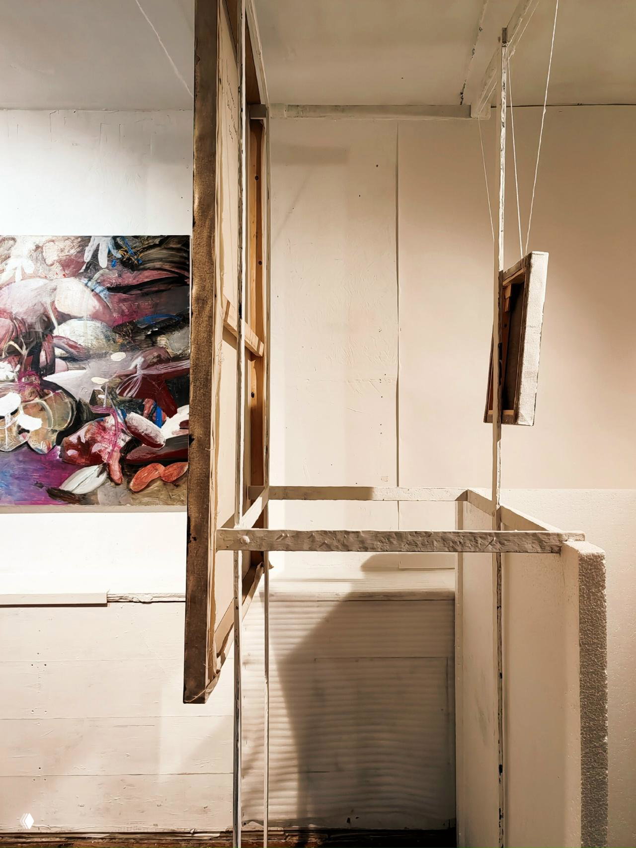 Вид с торца выставочной перегородки: оборотная сторона холста, деревянные рамы и канаты подвеса на фоне нейтральной белой стены галереи.