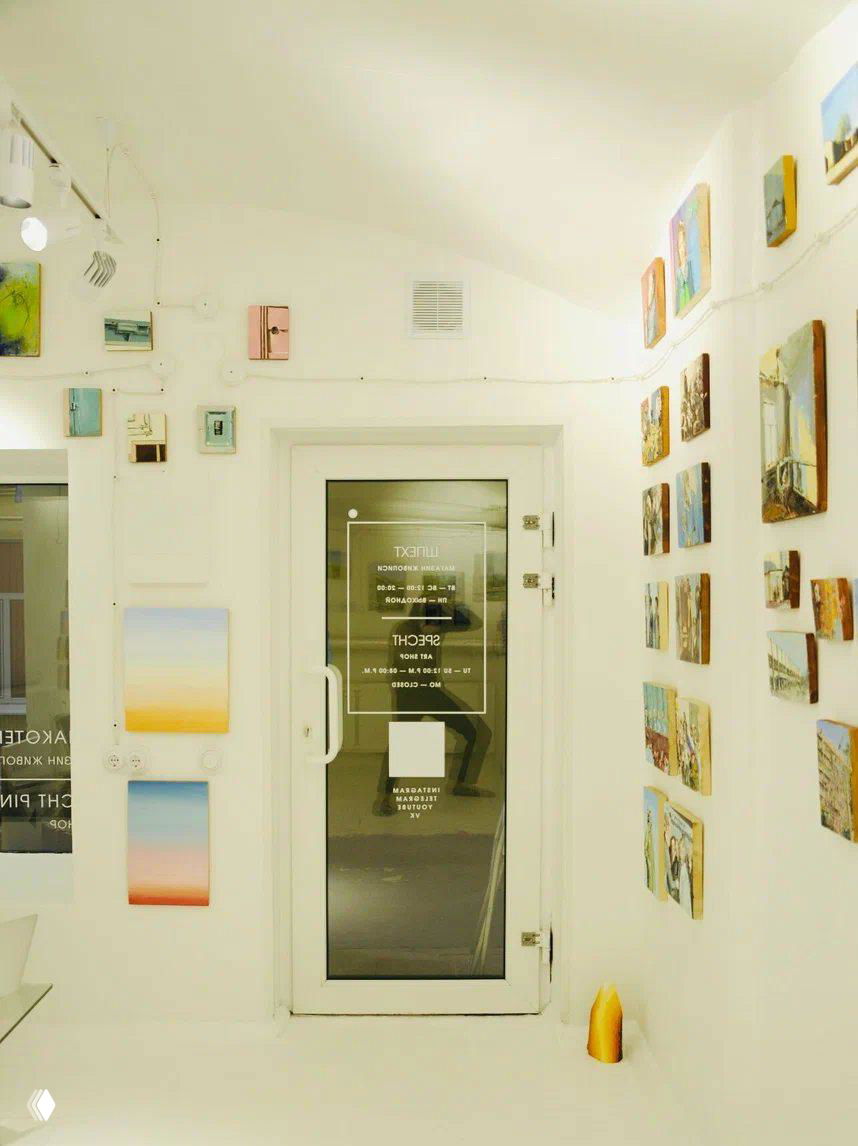 Входная зона галереи с белыми стенами и стеклянной дверью: на двери наклейки с названием и режимом работы, отражение интерьера внутри.